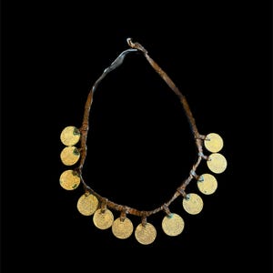 May include: A necklace with multiple gold-coloured, circular pendants. The pendants are attached to a twisted, brown cord. The necklace is set against a black background, highlighting the gold colour of the pendants.