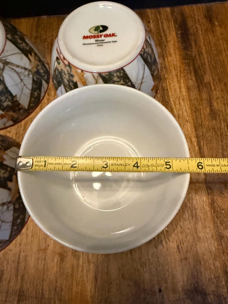 May include: A white ceramic bowl with a diameter of approximately 13 cm is measured with a tape measure. The bowl is part of a set with other bowls and mugs. The mugs have a camouflage pattern and the text "MOSSY OAK. Winter".