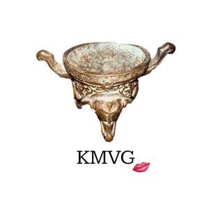 May include: A bronze-coloured, three-legged vessel with ornate detailing and two curved handles. The bowl-shaped top has a weathered appearance. The text "KMVG" is below the vessel, with a red lip graphic.