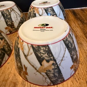 May include: Four white ceramic bowls with a camouflage design of brown and black tree branches and tan leaves. The bowls have a red rim and the text "Mossy Oak Winter" is printed on the base.