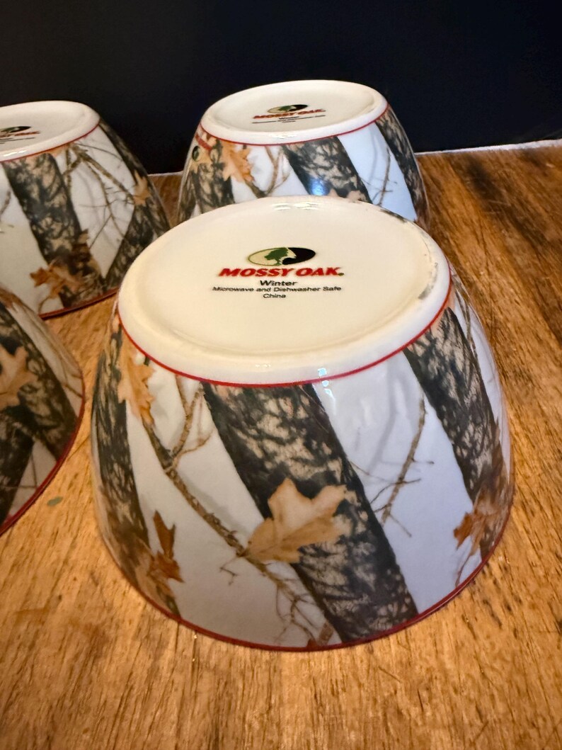 May include: Four white ceramic bowls with a camouflage design of brown and black tree branches and tan leaves. The bowls have a red rim and the text "Mossy Oak Winter" is printed on the base.