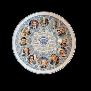 May include: A decorative ceramic plate with a blue and white floral border. The plate features portraits of historical figures around a central calendar design with the text "Millennium 2000 Art & Music".
