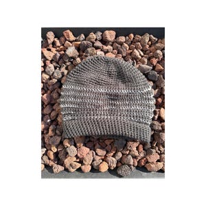 May include: A gray crocheted beanie hat with a ribbed texture and a white stripe detail. The hat is displayed on a bed of small, reddish-brown rocks. The hat appears to be handmade.