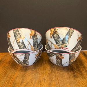 May include: Four white ceramic bowls with a camouflage pattern of brown, black, and tan. The bowls have a red rim and a dark interior. The bowls are stacked in pairs on a wooden surface.