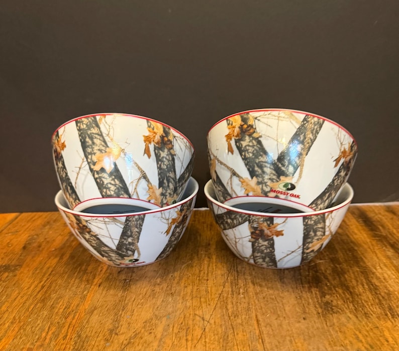 May include: Four white ceramic bowls with a camouflage pattern of brown, black, and tan. The bowls have a red rim and a dark interior. The bowls are stacked in pairs on a wooden surface.