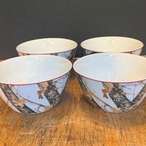 May include: Four white ceramic bowls with a camouflage design around the outside. The bowls have a red trim around the rim and are on a wooden surface.