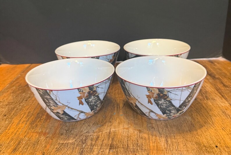 May include: Four white ceramic bowls with a camouflage design around the outside. The bowls have a red trim around the rim and are on a wooden surface.