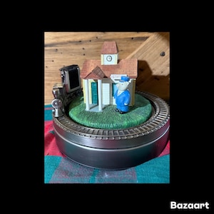Op de afbeelding: Een miniatuur treinstation scène met een klokkentoren, een kleine trein en een figuur in een blauw uniform. Het station heeft een bruin dak en een groene grasbasis, geplaatst op een cirkelvormig spoor. De basis is van zilverkleurig metaal.