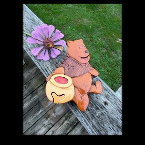 May include: A handcrafted wooden bear figurine featuring a glossy finish, depicted in a seated position while holding a light-colored wooden honey pot with a red center accent. The bear is constructed from various wood tones, showcasing a dark brown vest.