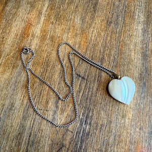 May include: A silver chain necklace with a heart-shaped pendant. The pendant is white with iridescent streaks. The necklace is lying on a wooden surface with a natural wood grain pattern.