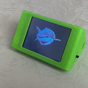 May include: A small, rectangular device with a bright green frame. The screen displays a detailed, blue illustration of a shark. The device is angled slightly, showcasing its design and the image on the screen.