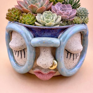 May include: A whimsical ceramic planter shaped like a face, with closed eyes and a gold nose ring. The planter is filled with a variety of succulents in shades of green, pink, and purple. The planter has a light blue rim and is set against a soft pink background.