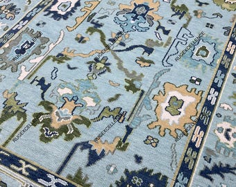 May include: A rectangular, light blue rug with an intricate floral pattern. The rug features a central field of light blue with various floral motifs in shades of green, beige, and navy blue. A navy blue border frames the rug, adding a contrasting element to the design.