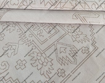 May include: Close-up of a beige rug with a geometric pattern in light brown. The rug has a detailed design with repeating shapes and lines, creating a textured appearance. The edges of the rug are visible, showing the layered construction.