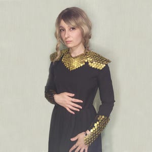 Könnte beinhalten: Ein schwarzes Kleid mit langen Ärmeln, das goldfarbene, schuppenartige Schulter- und Armakzente aufweist. Das Kleid hat ein schlichtes, elegantes Design, wobei die metallischen Akzente einen Hauch von Fantasie oder historischem Flair verleihen. Das Model trägt das Kleid.