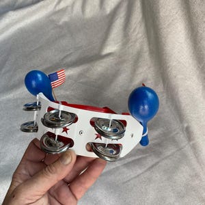 May include: A red, white, and blue toy tambourine with a small American flag. The tambourine has a white body with silver jingles and blue balloon-shaped handles. The design includes red stars.