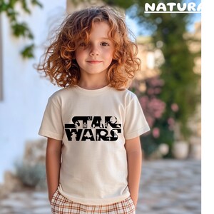 May include: A cream-colored t-shirt featuring the text "STAR WARS" in a black, stylized font. The shirt is worn by a child with curly red hair. The word "NATURA" is printed in green at the top right.