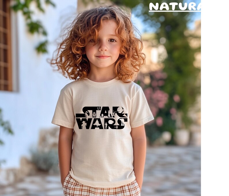 May include: A cream-colored t-shirt featuring the text "STAR WARS" in a black, stylized font. The shirt is worn by a child with curly red hair. The word "NATURA" is printed in green at the top right.