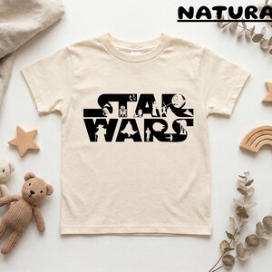 May include: Cream-colored t-shirt featuring the black text "STAR WARS" with character silhouettes. The shirt is surrounded by wooden stars, a wooden rainbow, and two teddy bears.