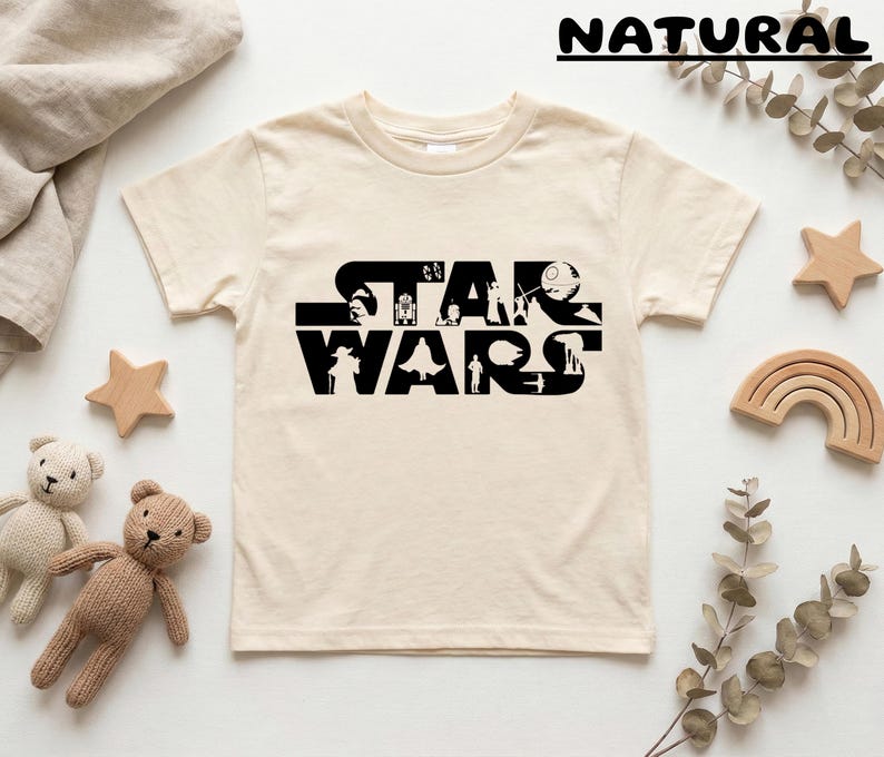 May include: Cream-colored t-shirt featuring the black text "STAR WARS" with character silhouettes. The shirt is surrounded by wooden stars, a wooden rainbow, and two teddy bears.