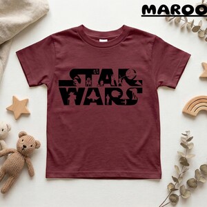 May include: Maroon t-shirt featuring the black text "STAR WARS" with character silhouettes. The shirt is surrounded by wooden stars, a rainbow, and teddy bears.
