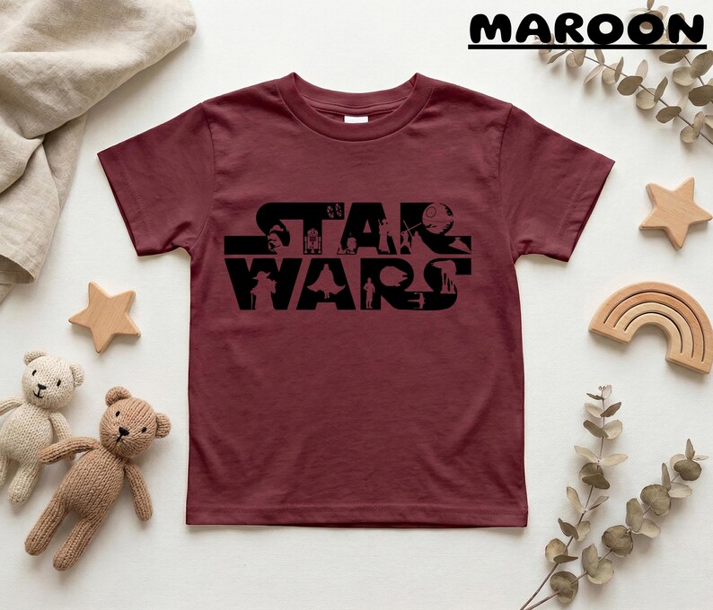 May include: Maroon t-shirt featuring the black text "STAR WARS" with character silhouettes. The shirt is surrounded by wooden stars, a rainbow, and teddy bears.