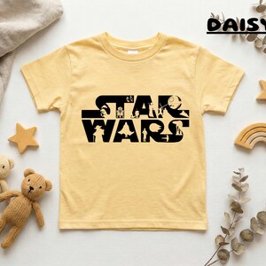 May include: A light yellow t-shirt featuring the black text "STAR WARS" with character silhouettes. The shirt is surrounded by decorative wooden stars, a rainbow, teddy bears, and greenery. The word "DAISY" is written in black script at the top right.