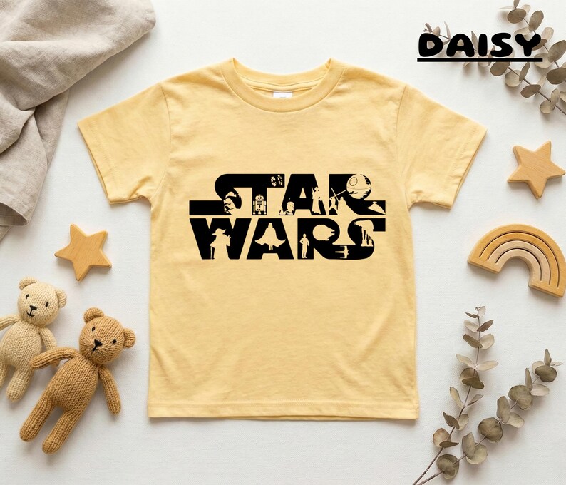 May include: A light yellow t-shirt featuring the black text "STAR WARS" with character silhouettes. The shirt is surrounded by decorative wooden stars, a rainbow, teddy bears, and greenery. The word "DAISY" is written in black script at the top right.