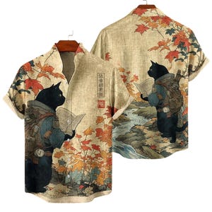 May include: Tan short-sleeve button-up shirt featuring a black cat wearing a backpack and reading a book, set against a backdrop of autumn leaves and a river. The shirt has a Japanese-inspired design with text on the front.
