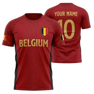 Op de afbeelding: Een bordeauxrood voetbalshirt met gouden letters en details. De voorkant toont "BELGIUM" en een schild met de Belgische vlag. De achterkant heeft "YOUR NAME" boven het nummer "10". Het shirt heeft zwarte zijpanelen.