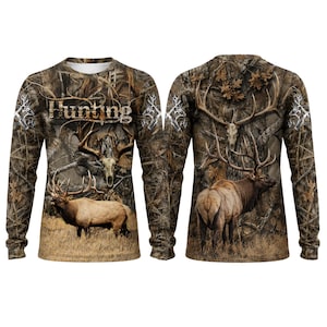 May include: A long-sleeve shirt featuring a camouflage pattern with forest imagery, including elk and deer skulls. The front displays the word Hunting in a stylized font. The design covers the entire garment, showcasing brown, tan, and green tones.
