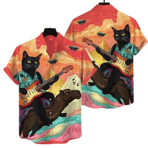 May include: Short-sleeved button-down shirt featuring a black cat playing a guitar while riding a camel. The shirt has a colorful desert scene with pyramids, UFOs, and musical notes. The cat wears a teal shirt. The shirt is red and orange.