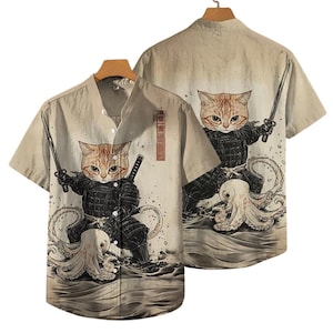Puede incluir: Camisa beige de manga corta con una ilustración de gato samurái. El gato, con armadura negra, empuña una espada y está sobre un pulpo. La camisa tiene cuello mandarín y cierre de botones. La parte trasera de la camisa también presenta la ilustración del gato.