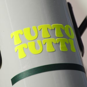 Könnte beinhalten: Nahaufnahme eines Fahrradrahmens in Hellgrau. Der Rahmen trägt die Worte "TUTTO TUTTI" in leuchtend gelben Buchstaben. Schwarze Kabel und ein dunkelgrüner Streifen sind ebenfalls sichtbar.