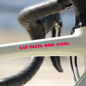 Könnte beinhalten: Nahaufnahme eines hellgrünen Fahrradrahmens mit schwarzen Lenkern und Akzenten. Der Rahmen trägt den Text "EAT PASTA RIDE FASTA" in leuchtend rosa Schrift. Das Fahrrad scheint für den Rennradsport konzipiert zu sein.