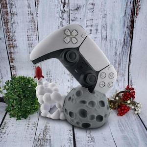 May include: A white and black game controller rests on a gray moon-shaped base with a rocket launching from a cloud. The controller has buttons and directional pads. A small red rocket is positioned above the cloud.