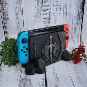 May include: A black Nintendo Switch console is displayed in a custom stand. The stand features a detailed, grey emblem design and is supported by black geometric shapes. The Switch has blue and red controllers.