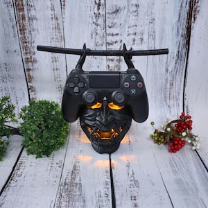 May include: A black gaming controller stand shaped like a demonic mask with glowing orange light emanating from the eyes and mouth. The stand features two black arms extending upwards to hold a black katana-style sword horizontally above the controller.