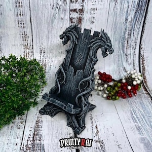 Könnte beinhalten: Ein grauer Handyhalter im Drachen-Design mit einem verwitterten, abgenutzten Aussehen. Der Ständer hat zwei Drachenköpfe an der Spitze und aufwendige Details. Auf der Basis steht der Text "PRINTY RAI 3D PRINT FOREST".