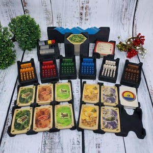 Könnte beinhalten: Ein Brettspiel-Organizer-Set mit verschiedenen Komponenten. Enthält schwarze Tabletts mit farbigen Spielsteinen und Kartenstapeln mit illustrierten Bildern. Würfel und andere Spielelemente sind ebenfalls sichtbar, alles angeordnet auf einer hellen Holzmaserungsoberfläche.