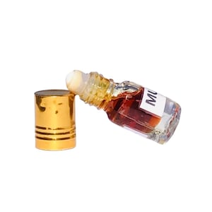 Könnte beinhalten: Kleine, klare Glasflasche mit Roll-on-Applikator, gefüllt mit einer dunkelbernsteinfarbenen Flüssigkeit. Die Flasche ist mit den Buchstaben "MU" beschriftet und liegt neben einem goldfarbenen Deckel mit horizontalen Rillen. Weißer Hintergrund.