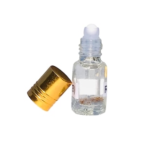 May include: A small, clear glass roller bottle with a white rollerball top and a gold cap. The bottle contains a clear liquid and a small amount of sediment. The gold cap is lying next to the bottle.