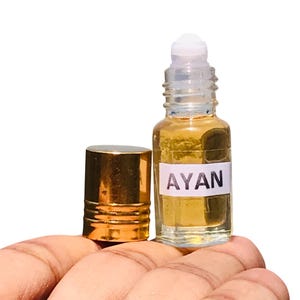 May include: A small, clear glass perfume bottle with a gold cap and a white label that reads "AYAN." The bottle contains a golden-colored liquid. The cap is off, and the bottle is held in a hand.