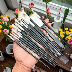 May include: A collection of artist paint brushes with black handles and various brush head shapes. The brushes are held in a hand, with a variety of flowers and plants in the background. The brushes are of different sizes and shapes, suitable for various painting techniques.