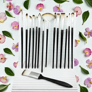 May include: A collection of artist paint brushes with black handles and white bristles, arranged on a white surface. The brushes vary in size and shape, including a fan brush. The background is decorated with pink flowers and green leaves.