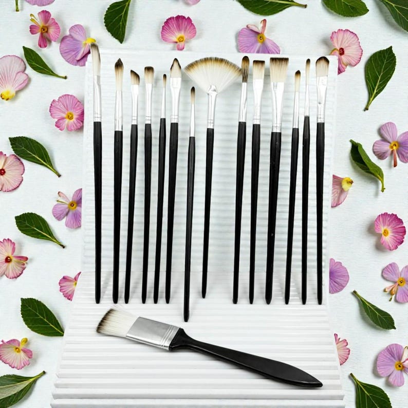 May include: A collection of artist paint brushes with black handles and white bristles, arranged on a white surface. The brushes vary in size and shape, including a fan brush. The background is decorated with pink flowers and green leaves.