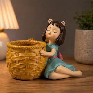 May include: A decorative figurine of a girl in a blue dress, embracing a woven basket-shaped planter. The planter is yellow and has a textured surface. The girl has brown hair and is wearing a red bow. The figurine is on a wooden surface.