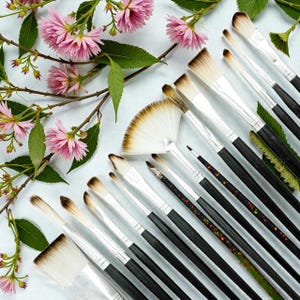 May include: An assortment of artist paint brushes with black handles and silver ferrules, arranged with pink flowers and green leaves on a light blue background. The brushes vary in size and shape, including a fan brush.