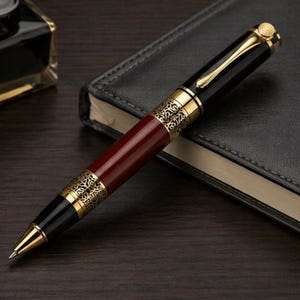 Peut inclure: Un stylo noir et doré avec un corps bordeaux repose sur un carnet noir en cuir. Le stylo présente des filigranes dorés et une pointe dorée. Un flacon d'encre est visible en arrière-plan.