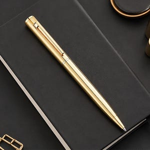 Peut inclure: Un stylo doré avec un clip et une pointe repose sur un cahier noir. Le stylo a une finition métallique brillante et un design épuré. C'est un instrument d'écriture.